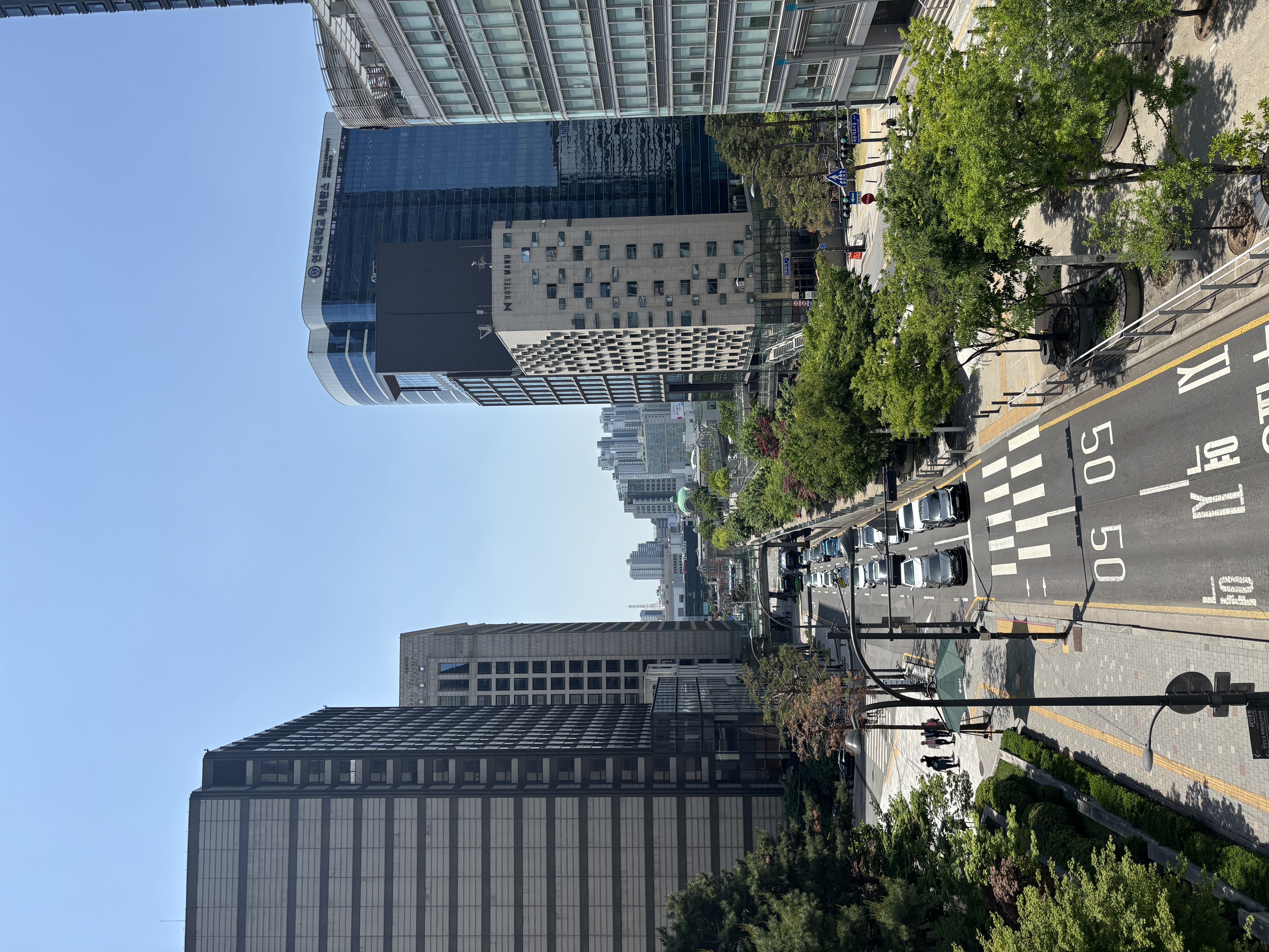 A view of a busy urban street lined with tall buildings and trees under a clear blue sky.