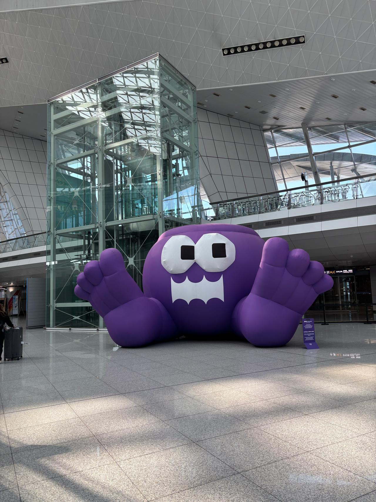 A large purple inflatable character with a smiling face and outstretched hands, situated in a bright, modern airport terminal.