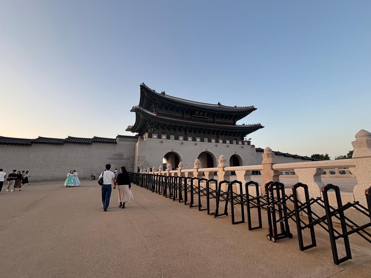 A couple walking towards the Gyeongbokgung Palace in Seoul, South Korea, with traditional architecture in the background and barriers lining the path.
