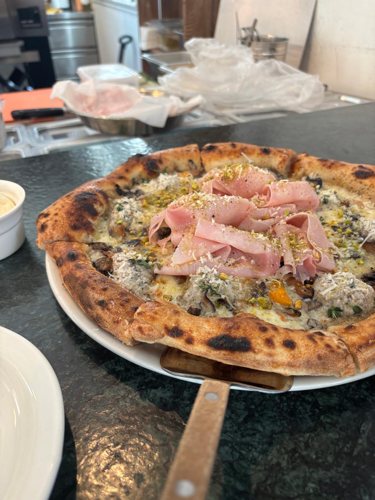 A freshly baked pizza topped with slices of ham, herbs, and cheese, served on a wooden peel with a dark stone countertop in the background.