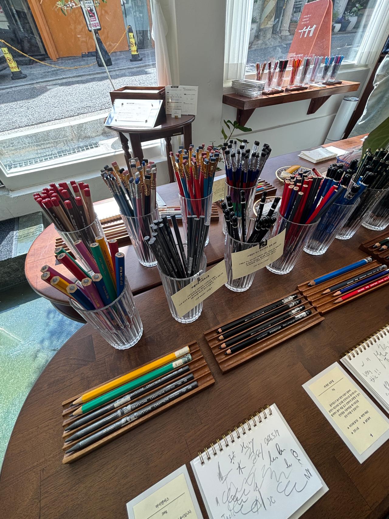 A display of various pens and pencils in clear containers on a wooden table, with notebooks and handwritten notes nearby, set in a well-lit space with large windows.