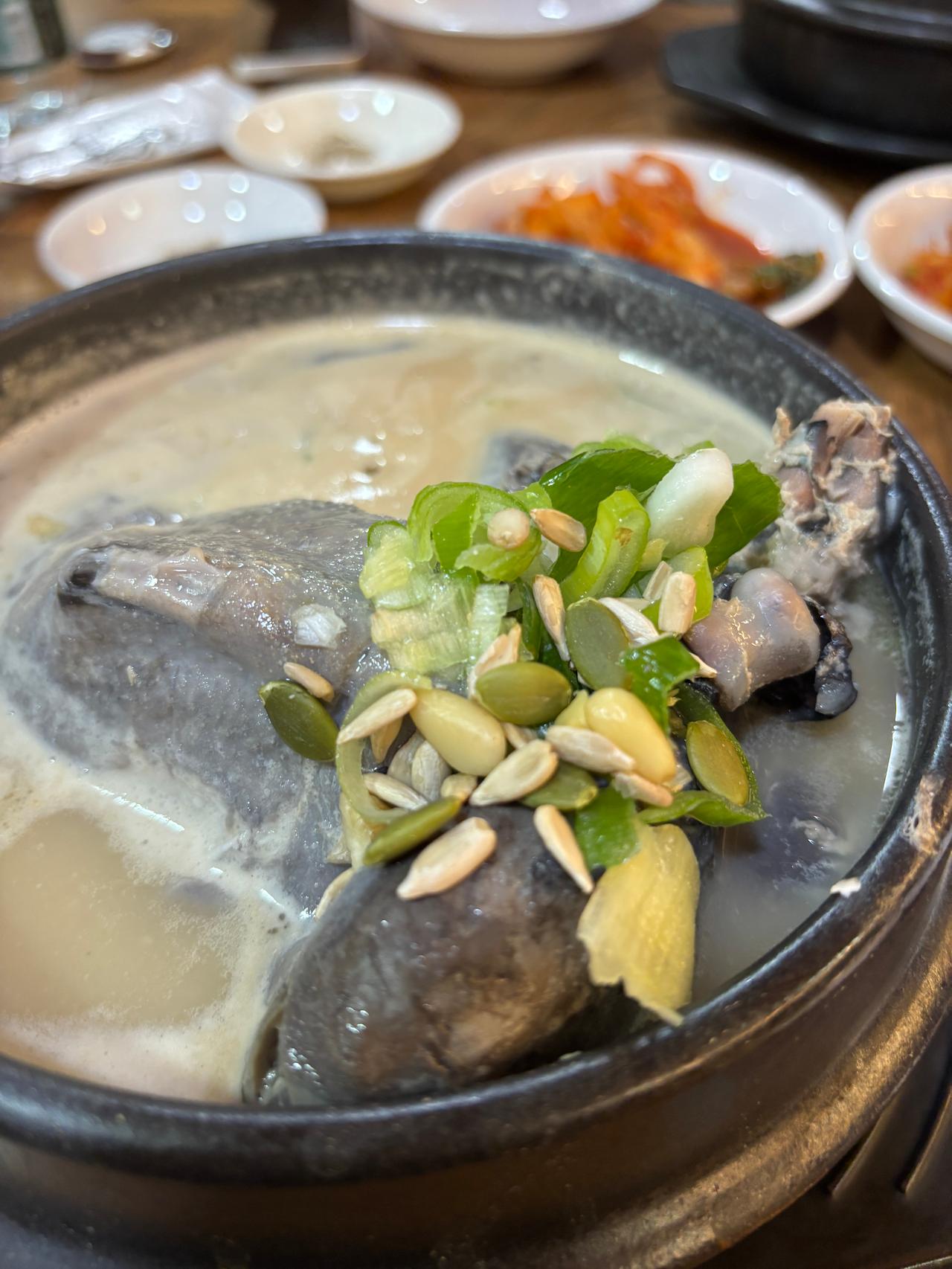 A close-up of a traditional Korean dish, showcasing a bowl of creamy soup with a whole chicken, garnished with green onions and seeds, surrounded by small side dishes.