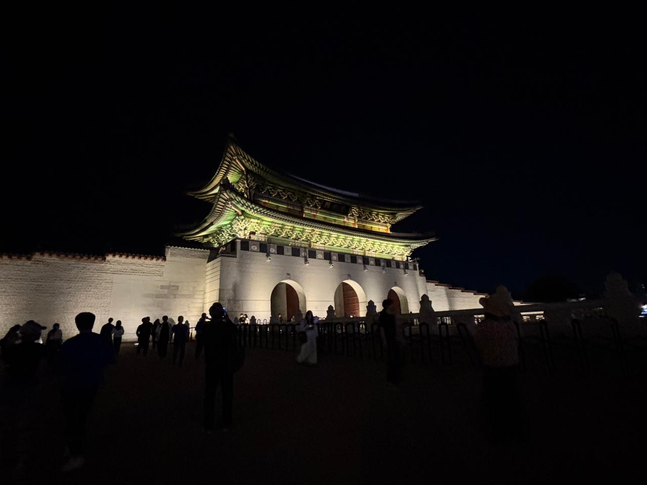 Night view of a historic building with a traditional Asian architectural style, illuminated against a dark sky, surrounded by visitors.