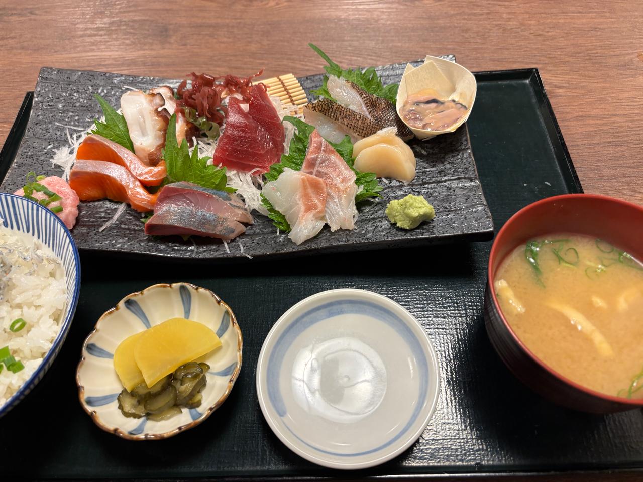 A beautifully arranged sashimi platter featuring various types of raw fish, garnished with leafy greens and wasabi, accompanied by a bowl of miso soup, pickled vegetables, and steamed rice in a patterned bowl.