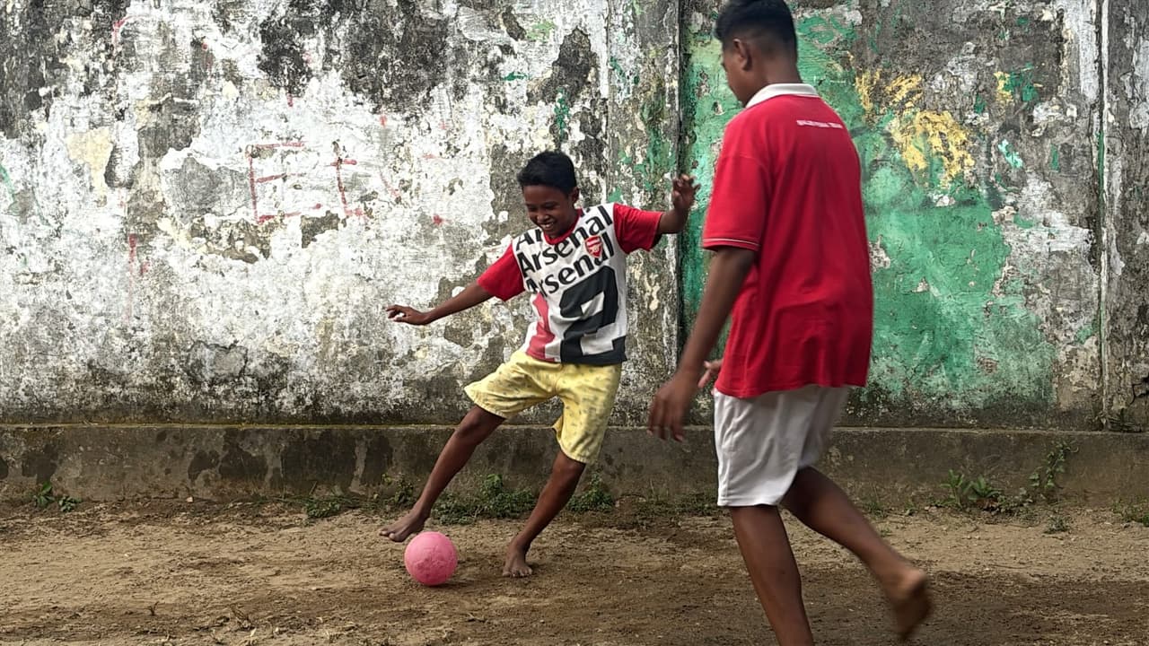 Local kids playing barefoot soccer with a pink ball against a weathered wall