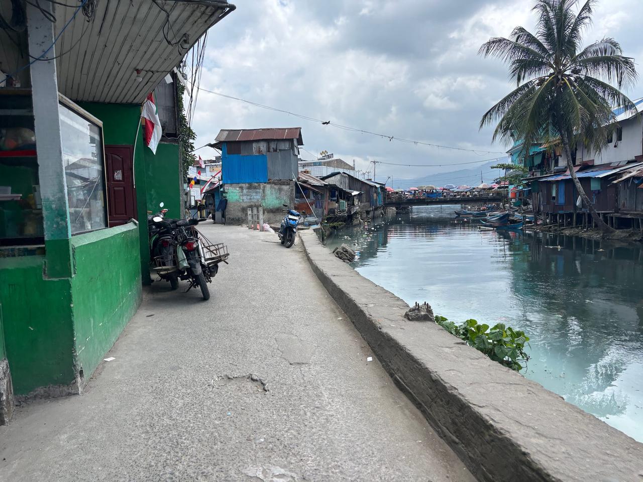 A chaotic backstreet of stilt houses and motorbikes reflected in still water
