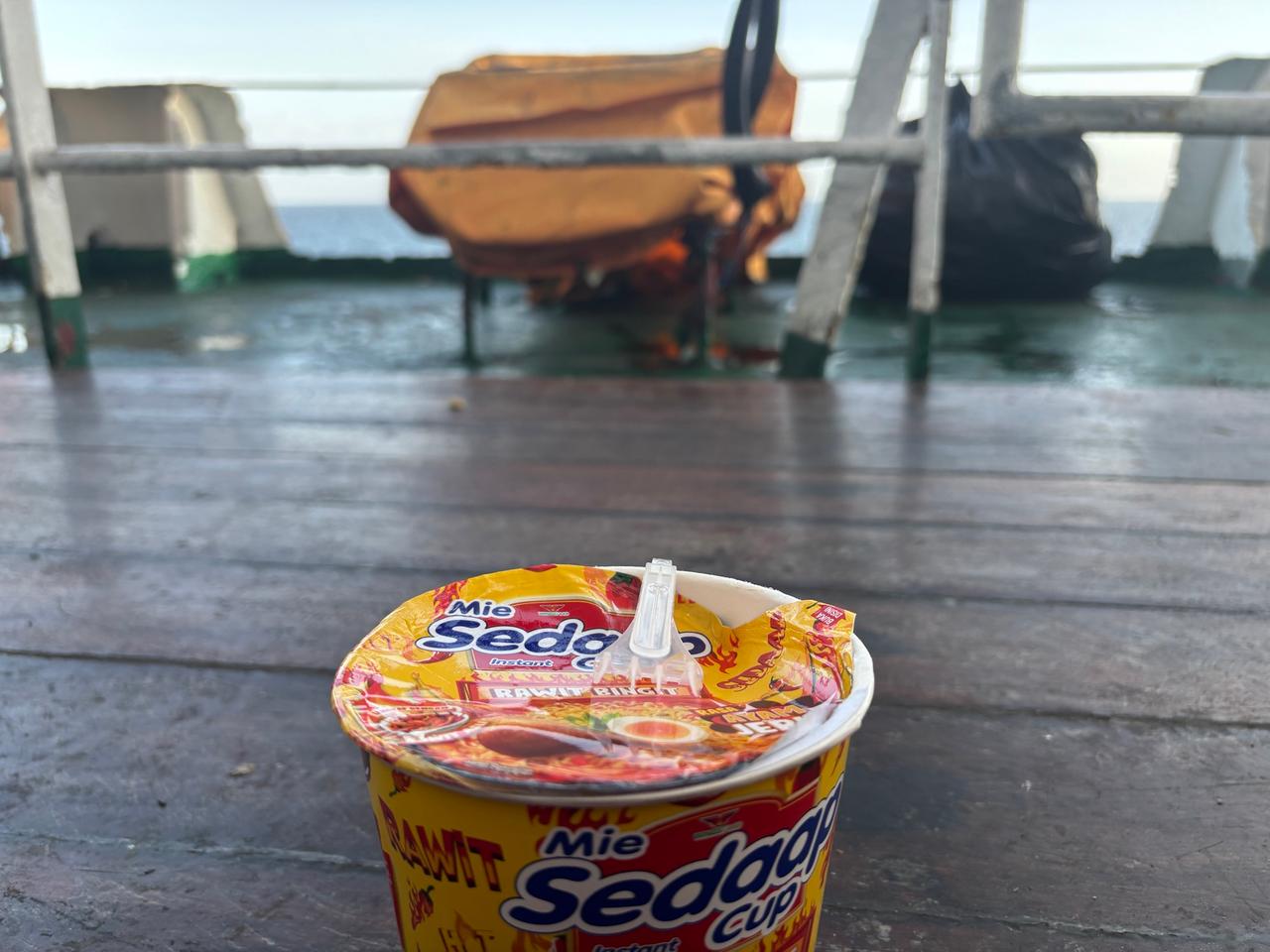 Semangkuk Pop Mie dengan pemandangan geladak feri di latar belakang