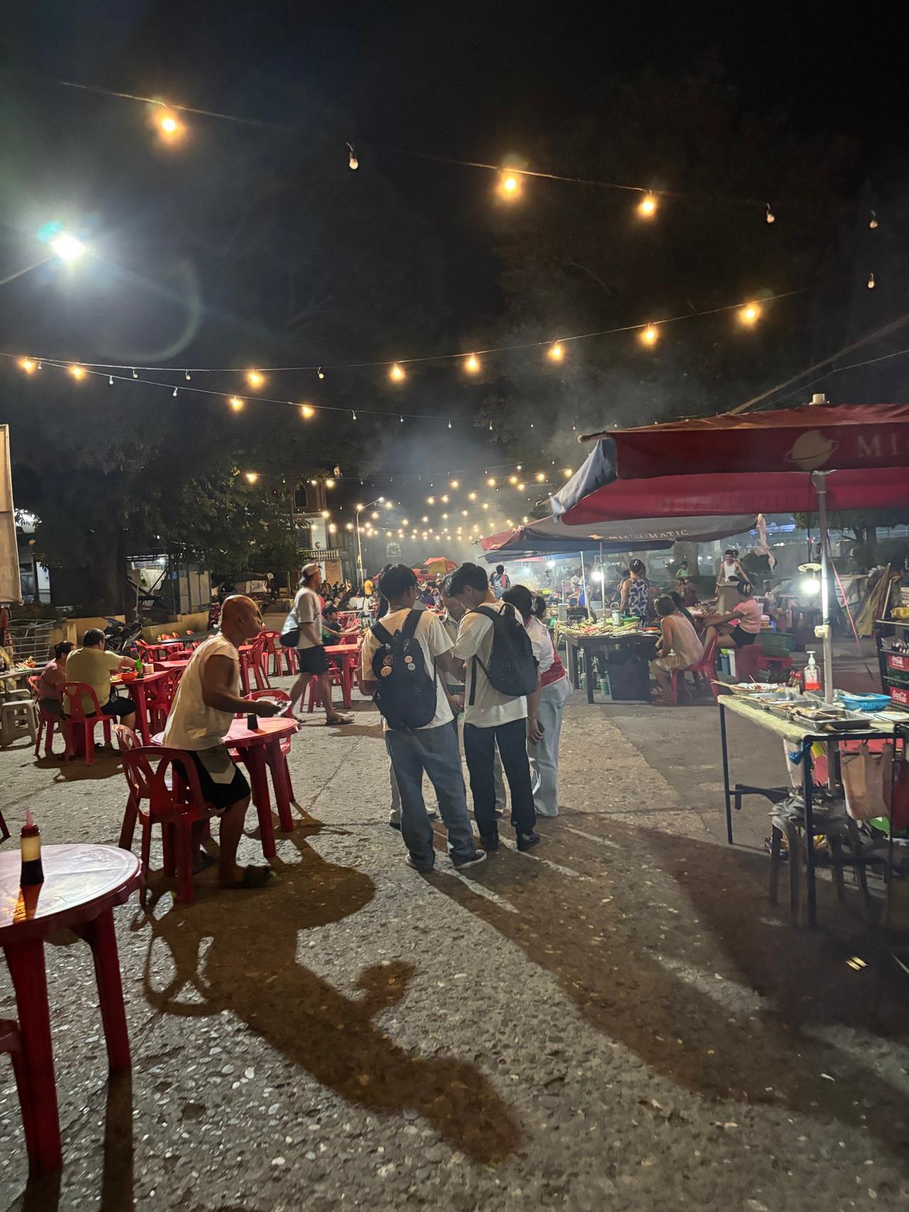 A lively outdoor street BBQ scene at night with string lights and people gathered around tables
