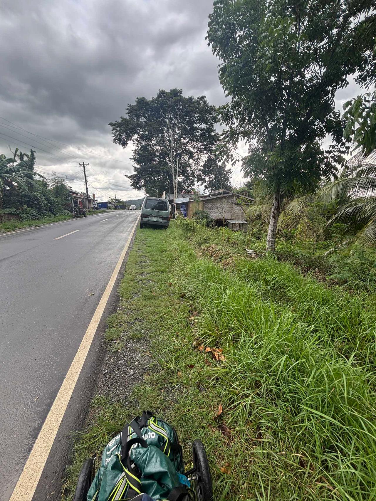 A quiet road under a cloudy sky with a travel cart parked on the grassy shoulder