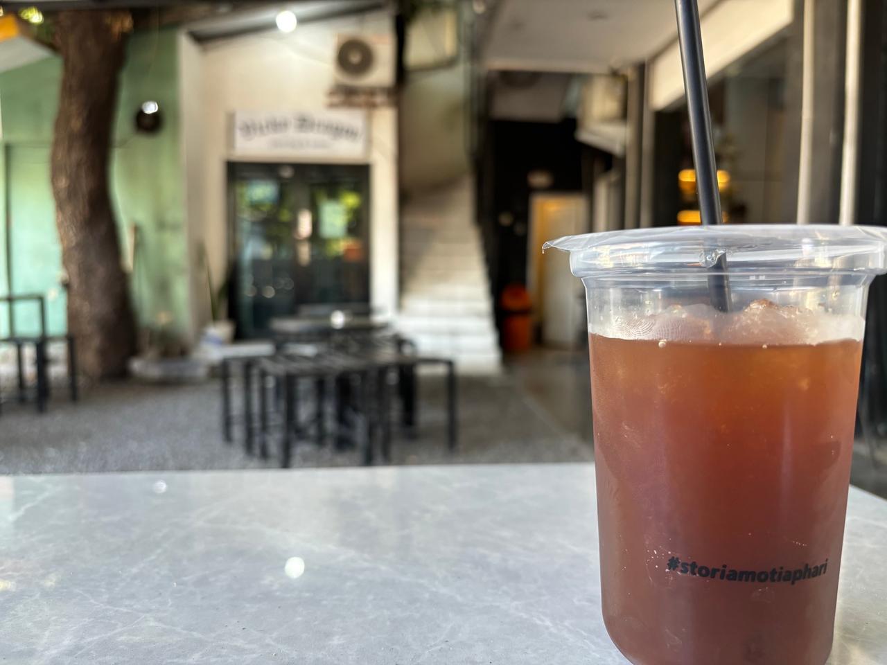 Segelas es kopi di atas meja kafe bertekstur marmer dengan halaman terbuka dan tangga di latar belakang