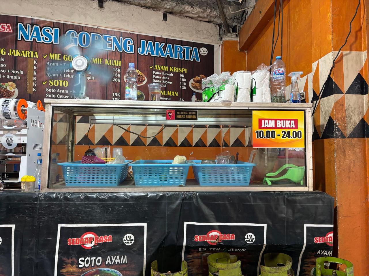 Papan menu warung makan lokal yang mencantumkan harga Mie Goreng, Mie Kuah, dan Soto di samping etalase kaca berisi makanan