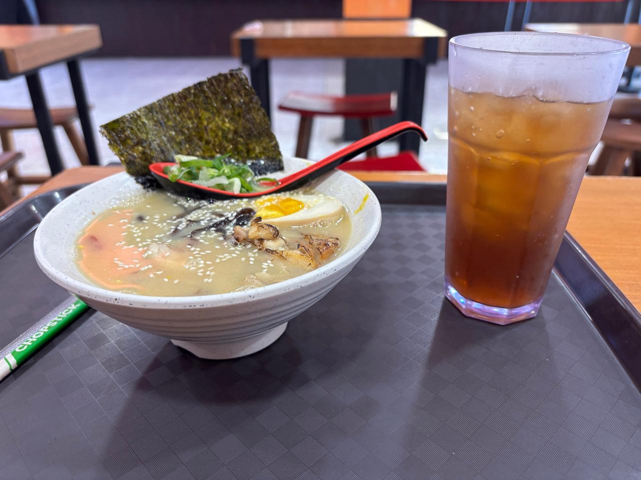 Semangkuk ramen creamy dengan nori, telur, dan ayam panggang di samping segelas es teh leci