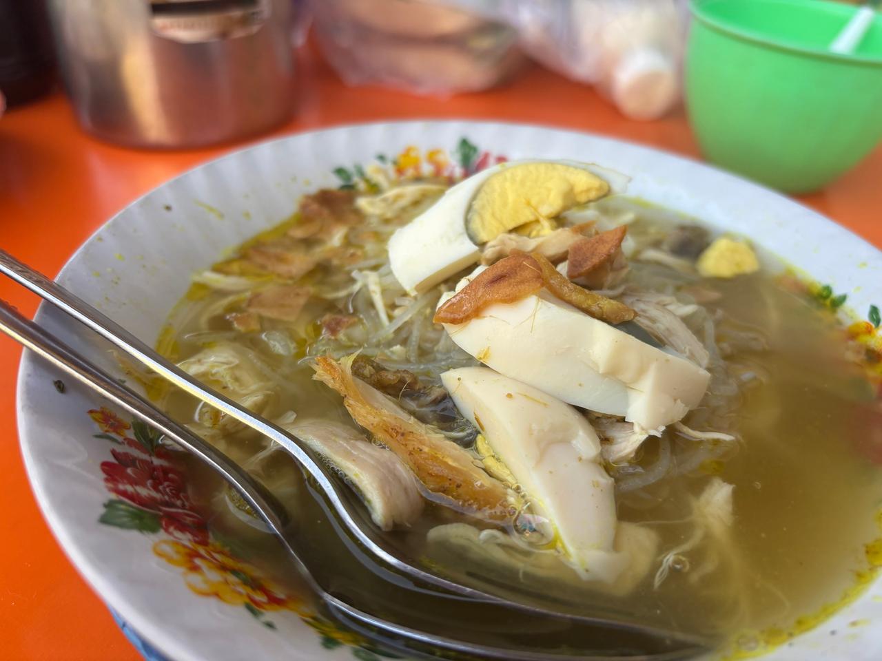 A bowl of yellow-broth chicken soup with sliced boiled egg, bean sprouts and fried shallots, with a spoon and fork resting in it