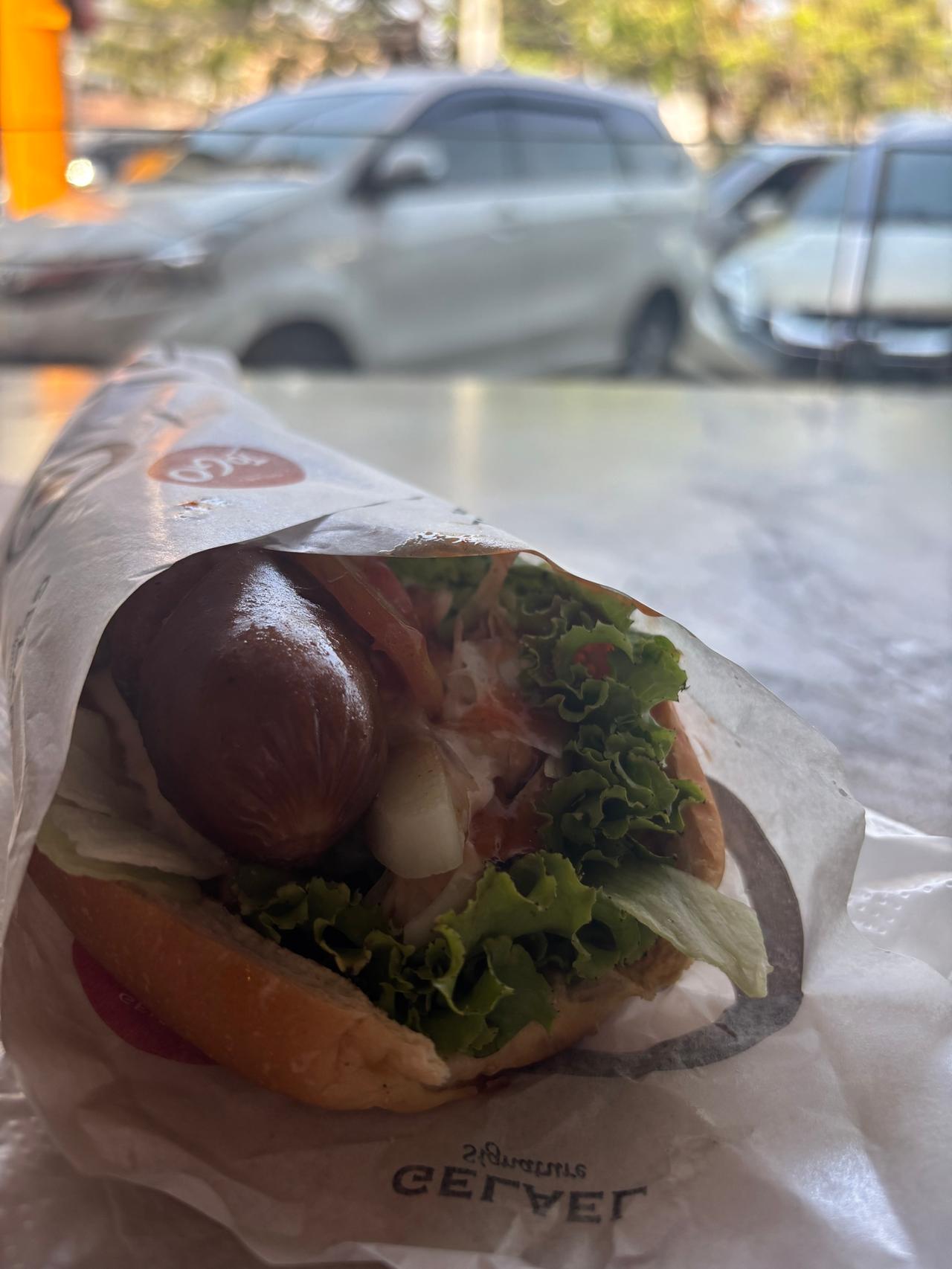 A hot dog with a glossy sausage, lettuce and onion wrapped in paper, with parked cars blurred behind it