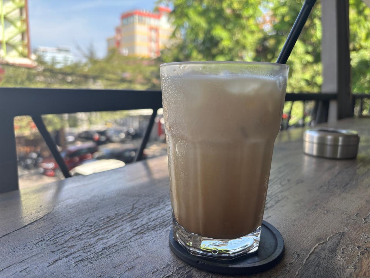 An iced milk coffee in a tall glass on a wooden table at an open-air cafe overlooking the street