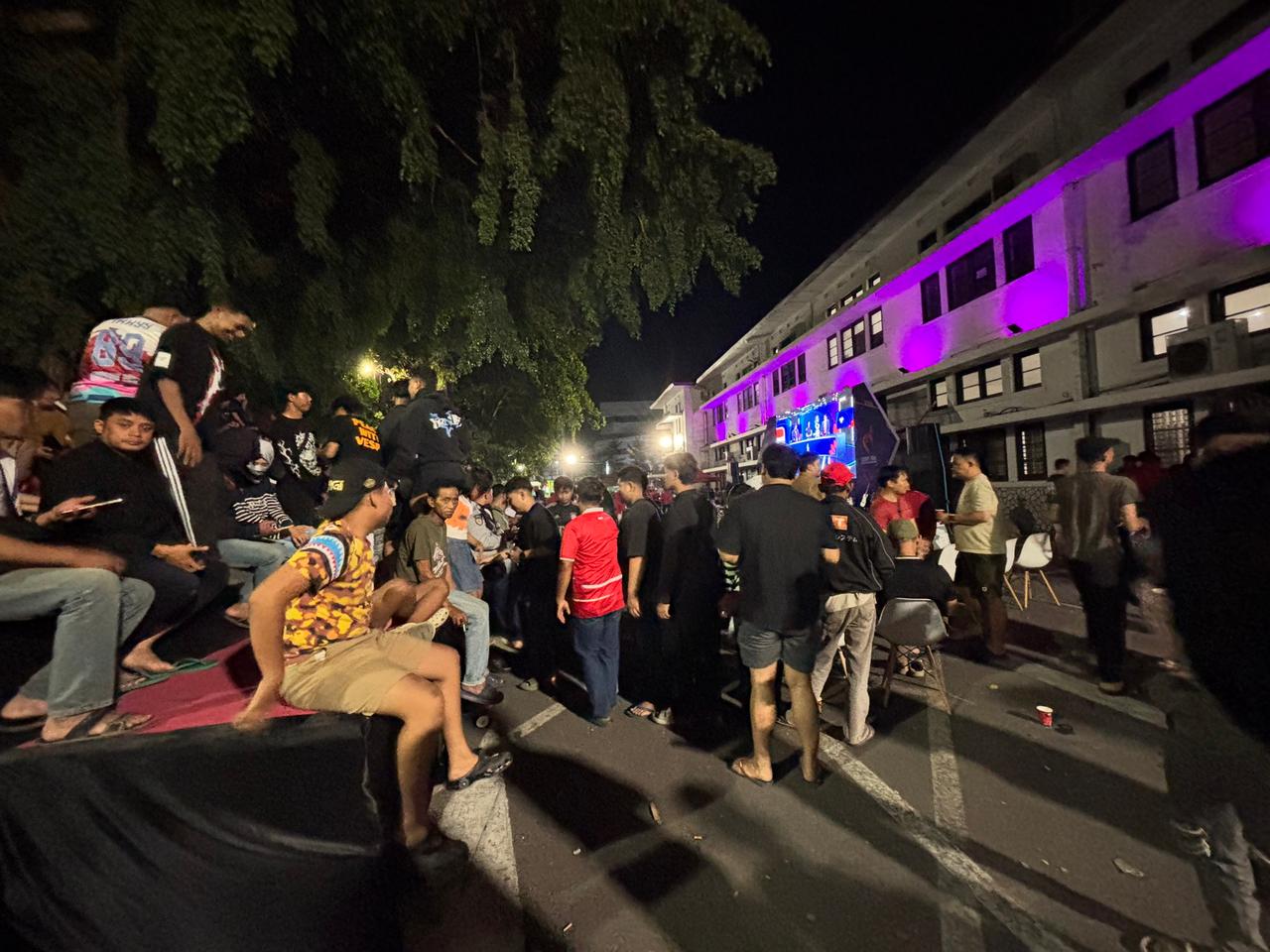 Crowds of fans gathered at night in the plaza in front of a purple-lit building, watching the match on a large screen