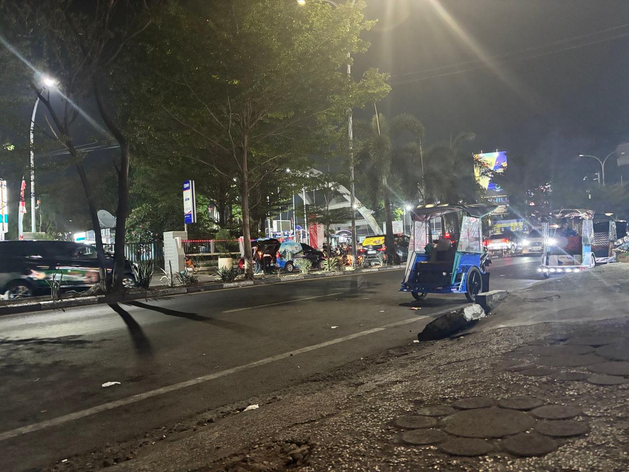 Sebuah jalan Makassar di malam hari dengan becak biru, mobil-mobil yang lewat, dan lampu-lampu jalanan yang sibuk