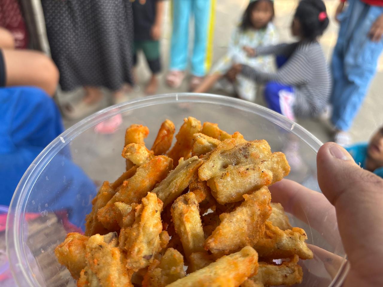 Sebuah wadah berisi camilan Tela-Tela yang baru digoreng, dipegang dengan anak-anak desa di latar belakang
