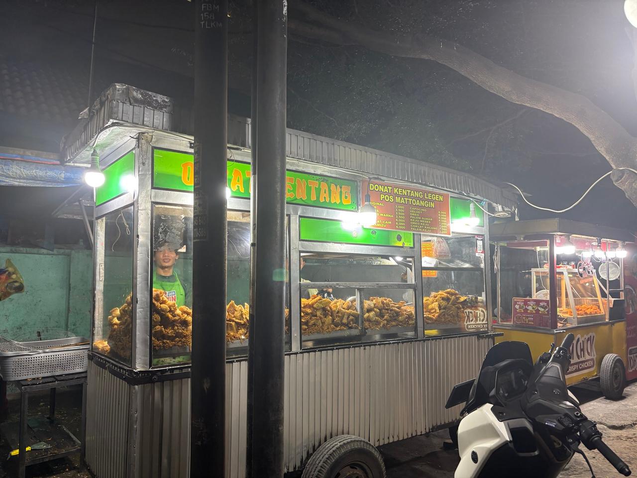 Sebuah lapak donat dan camilan gorengan yang terang benderang di malam hari dengan penjual yang terlihat di dalamnya