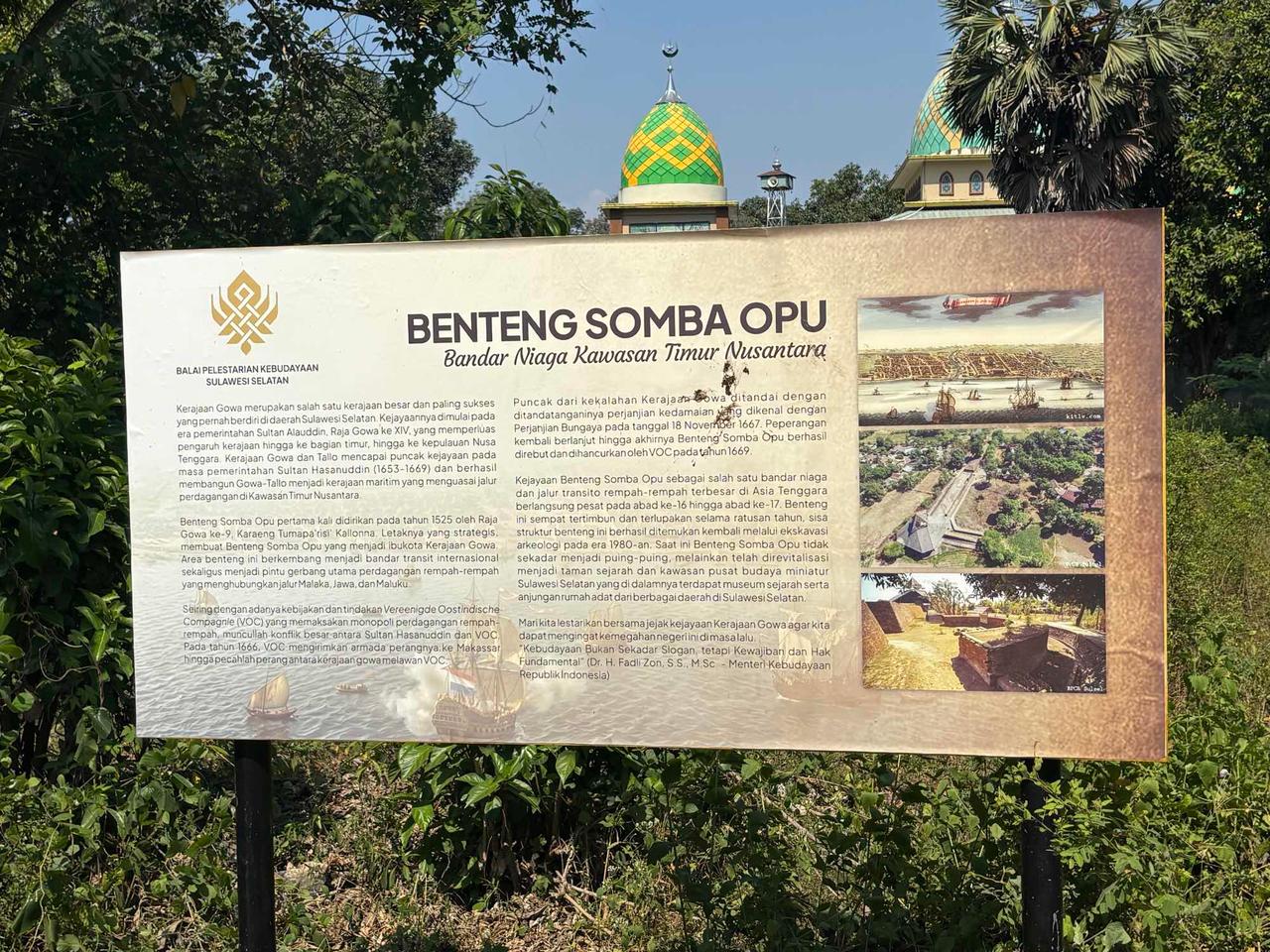 Papan informasi untuk Benteng Somba Opu dengan sebuah masjid berkubah hijau terlihat di balik pepohonan