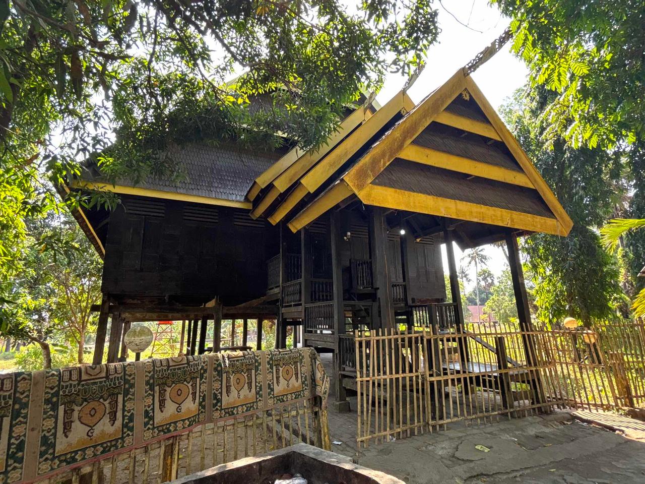 Sebuah rumah panggung kayu tradisional berwarna hitam dengan aksen kuning di dalam area Benteng Somba Opu
