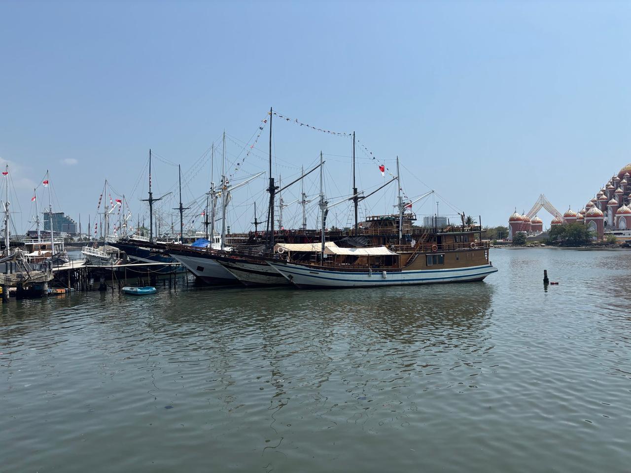 Kapal-kapal pinisi kayu tradisional yang berlabuh di pelabuhan Makassar dengan bangunan berkubah di latar belakang