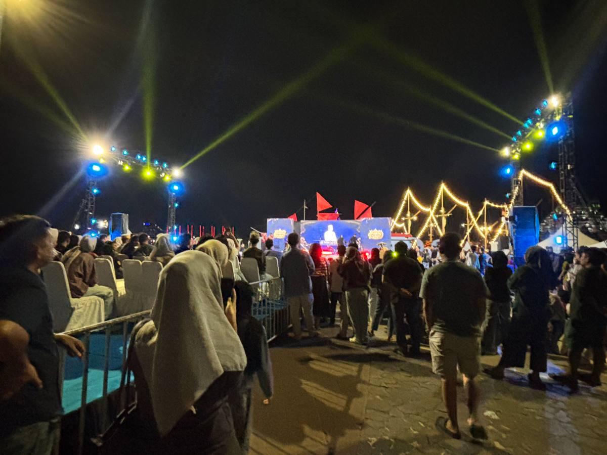 A crowd gathered at a nighttime festival on the waterfront, stage lights sweeping overhead