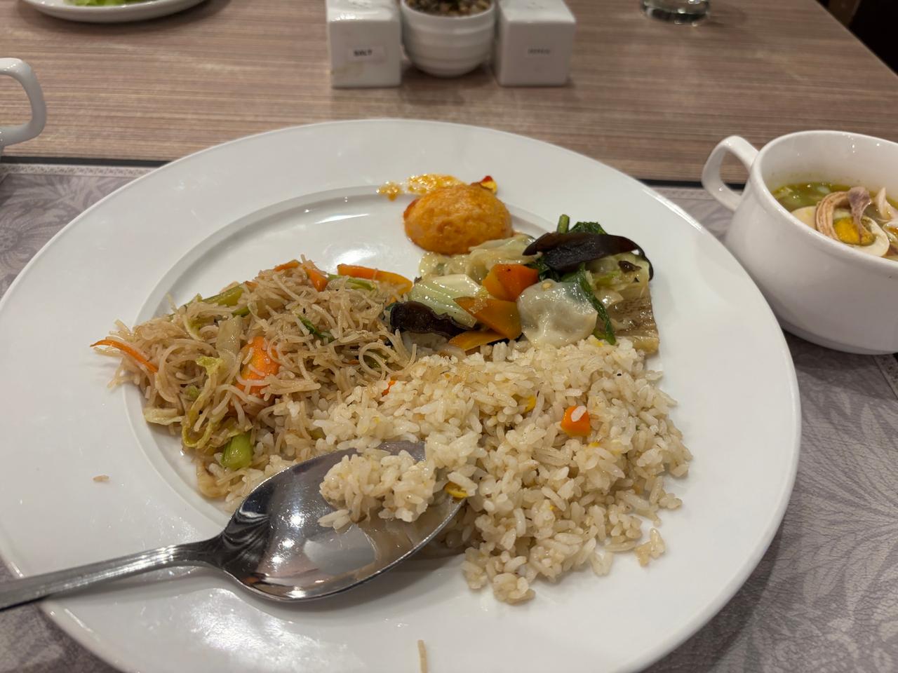 A hotel breakfast buffet plate with fried rice, noodles, vegetables and a side of soup