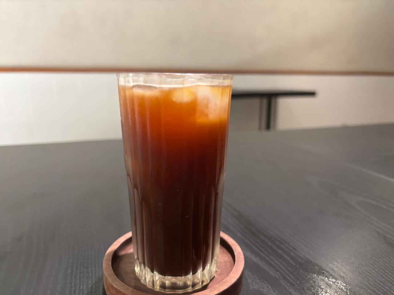 A tall glass of iced Americano coffee resting on a wooden coaster at a cafe table