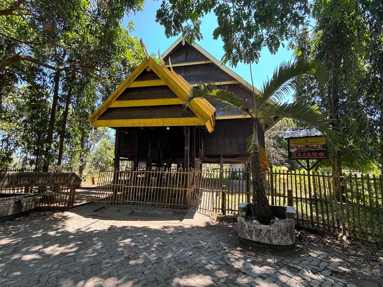 A traditional wooden "Rumah Adat" stilt house with a yellow-trimmed roof, framed by palm trees at Somba Opu