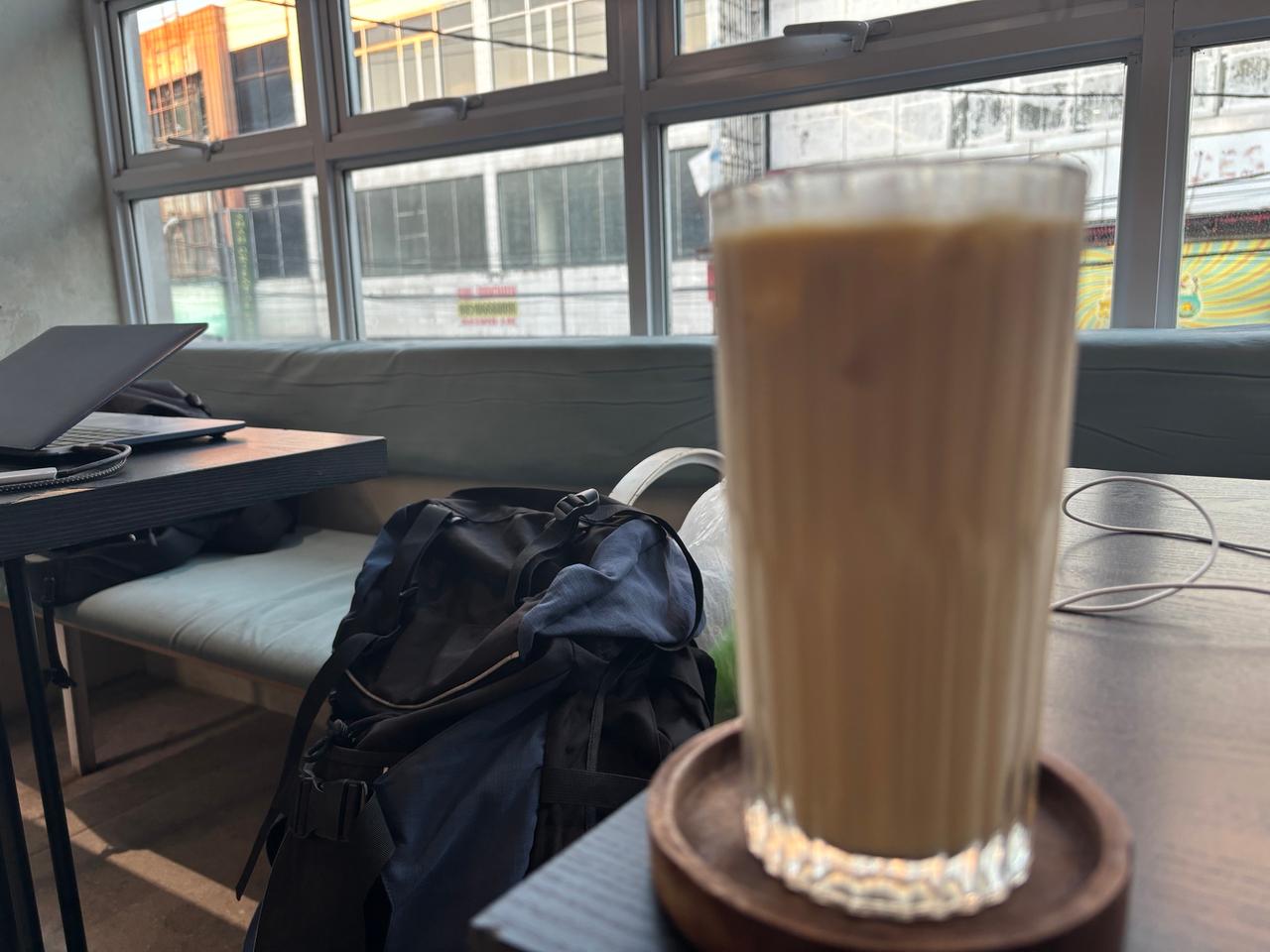 A backpack and iced coffee on a cafe table by a window, laptop open on a nearby bench in the evening light