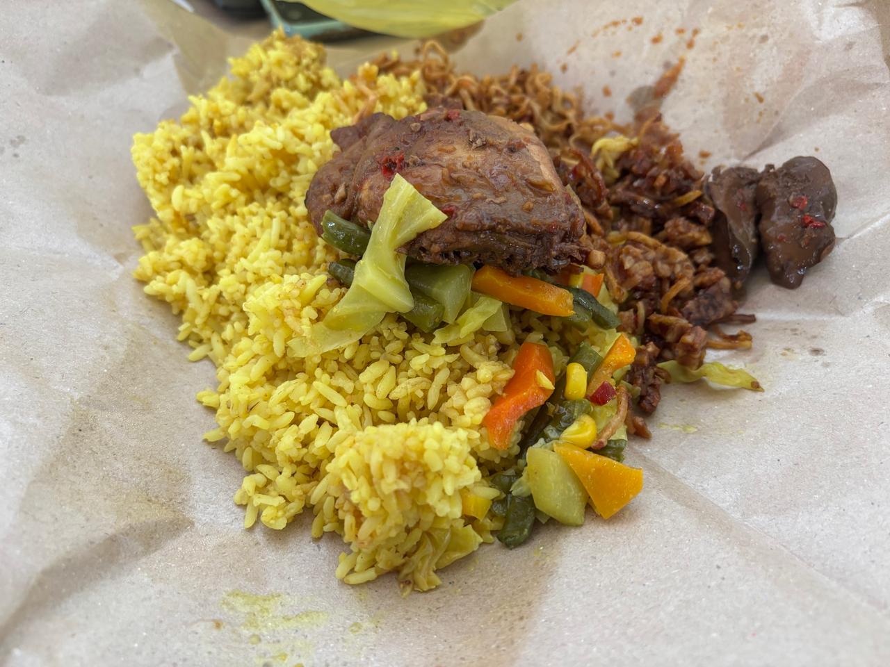 A street food meal of yellow rice, grilled chicken in sweet kecap manis, vegetables and crispy noodles wrapped in paper
