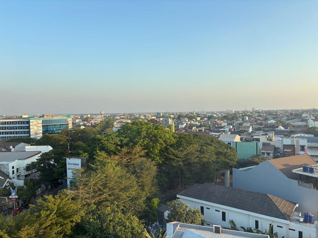 A hazy morning skyline view of Makassar city seen from a high floor, with dense trees and rooftops stretching to the horizon