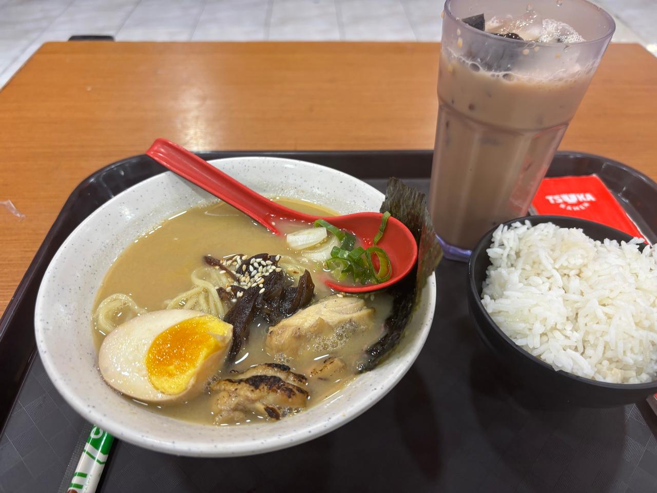 A bowl of ramen with a soft-boiled egg and grilled chicken, served with a side of rice and iced lychee tea