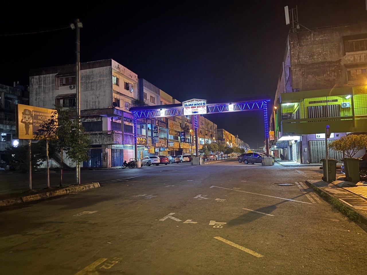 A quiet, empty street in Tawau at night under a lit welcome gate