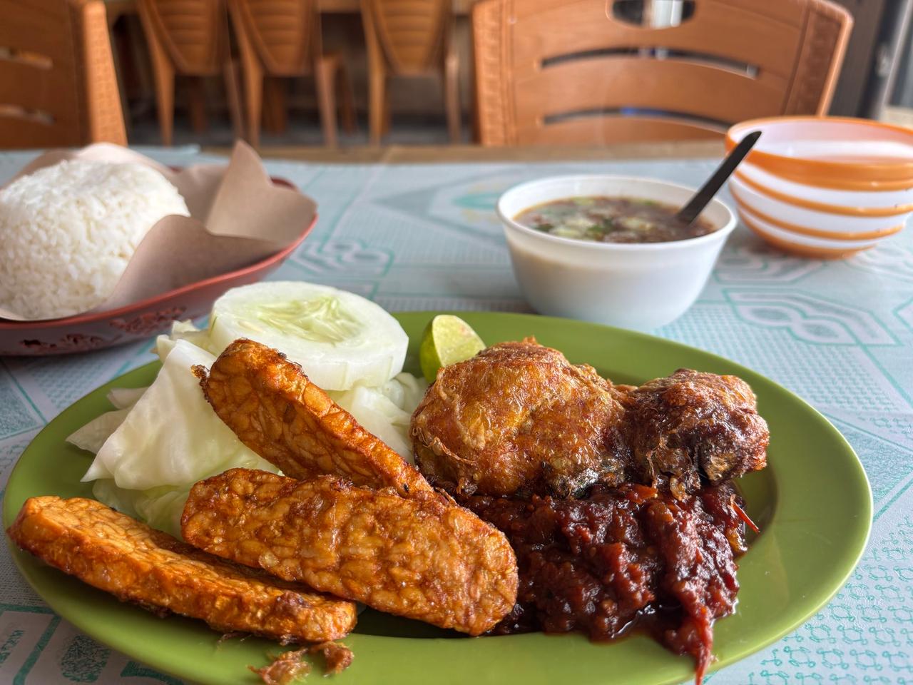 A plate of Lalapan with fried tempeh, fried chicken, sambal, cucumber and cabbage alongside rice and broth