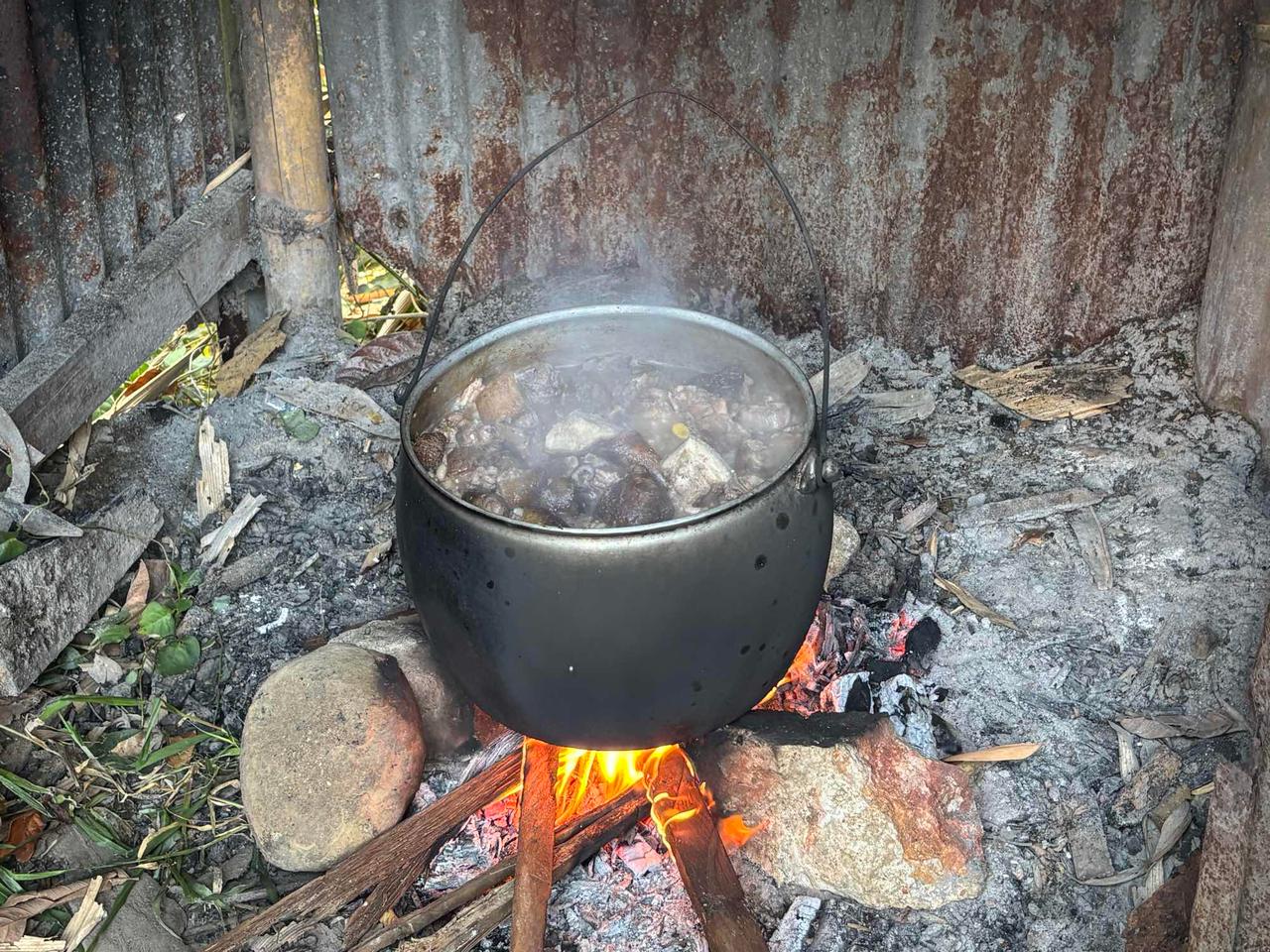 A blackened pot of pork broth simmering over an open fire, steam rising from the meat inside