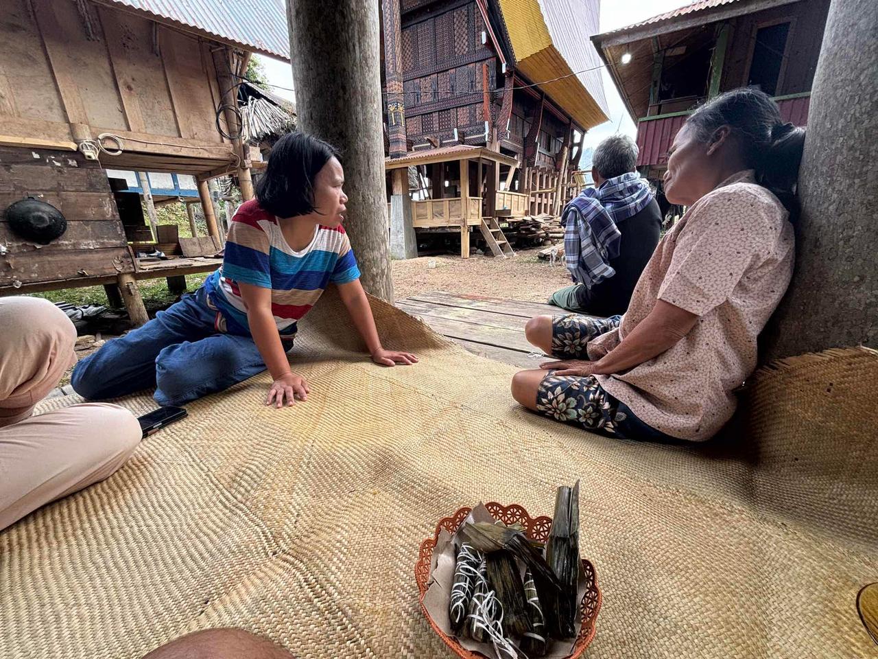 Sitting on a woven mat inside a traditional Tongkonan house, talking with an elderly local woman