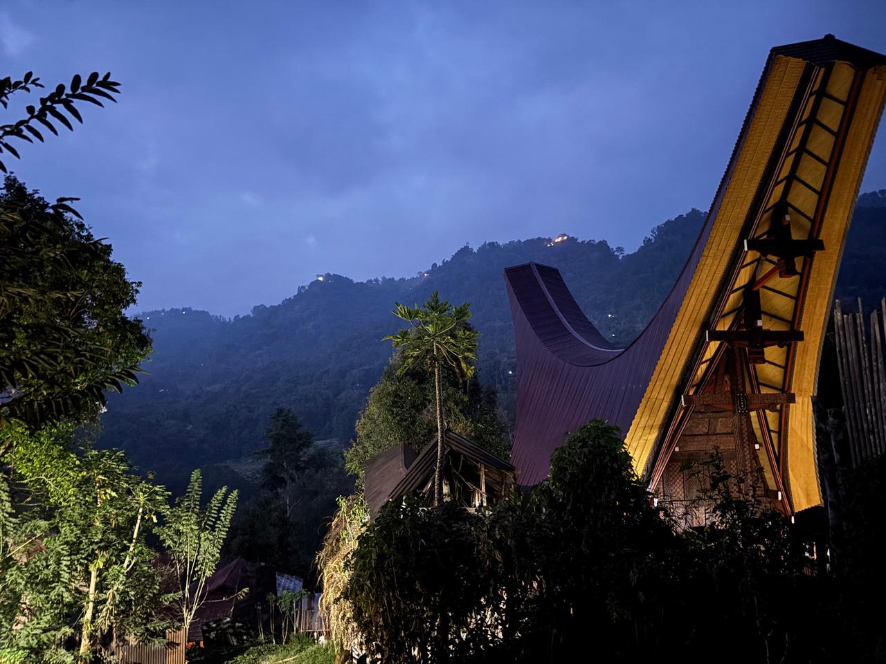 A Tongkonan house with its curved saddleback roof lit up against the pre-dawn mountain sky