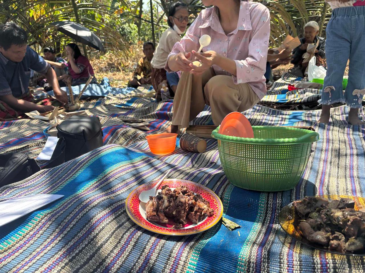 Villagers seated on mats under the trees sharing plates of cooked meat during the auction