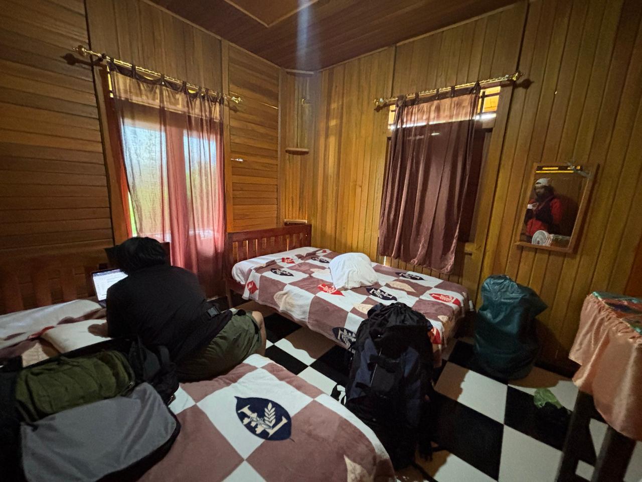 A wooden guesthouse room with backpacks and a checkered floor, a guest working on a laptop by the bed