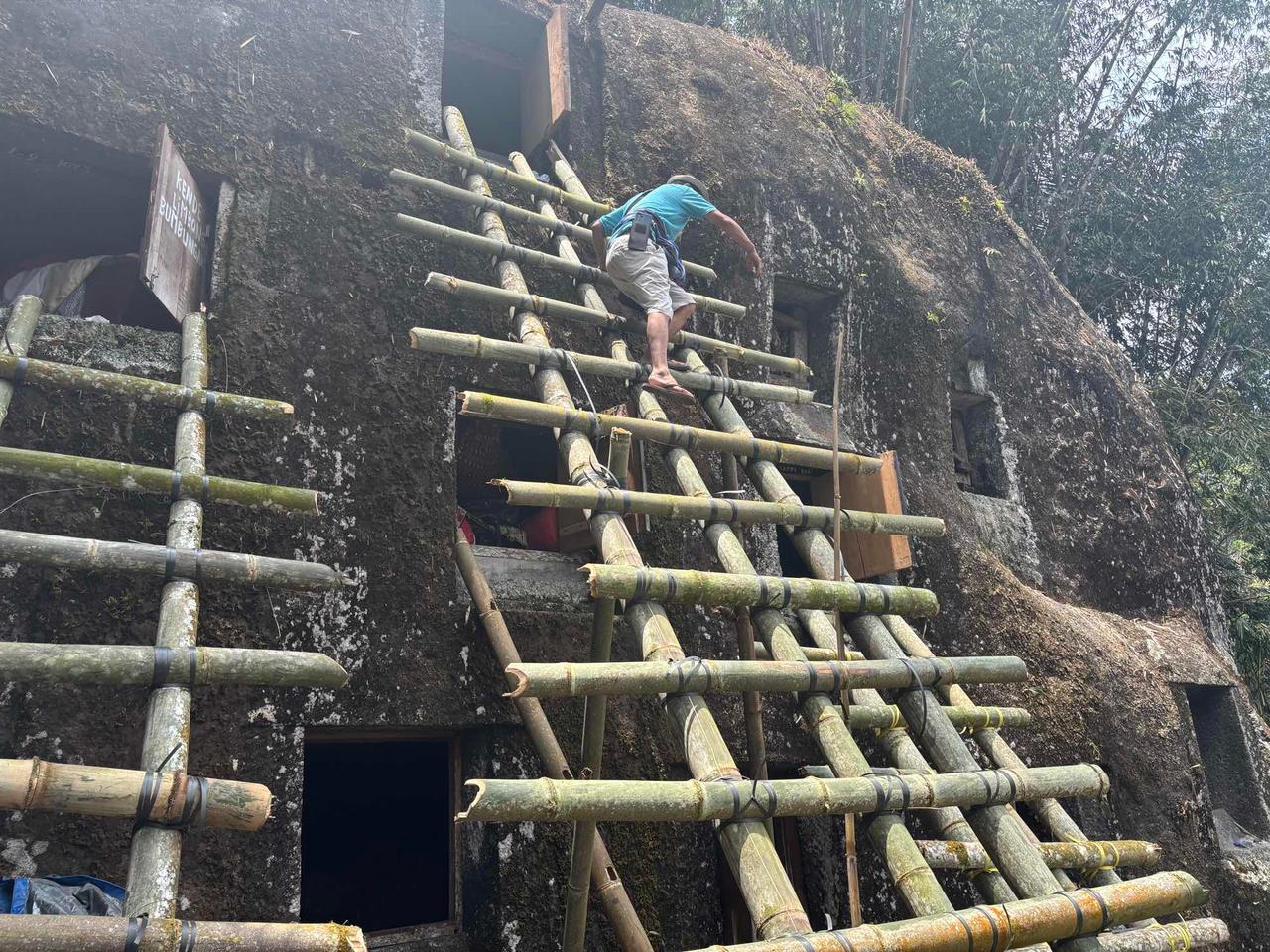 A man climbing a bamboo scaffold built against a moss-covered rock tomb to reach a burial niche