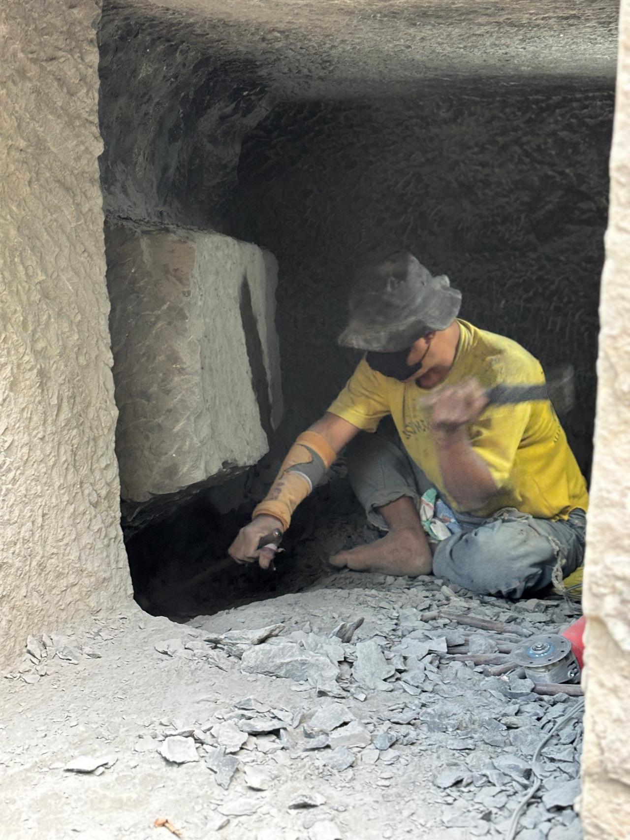 A worker carving open a stone burial niche inside a rock tomb, surrounded by chipped stone debris