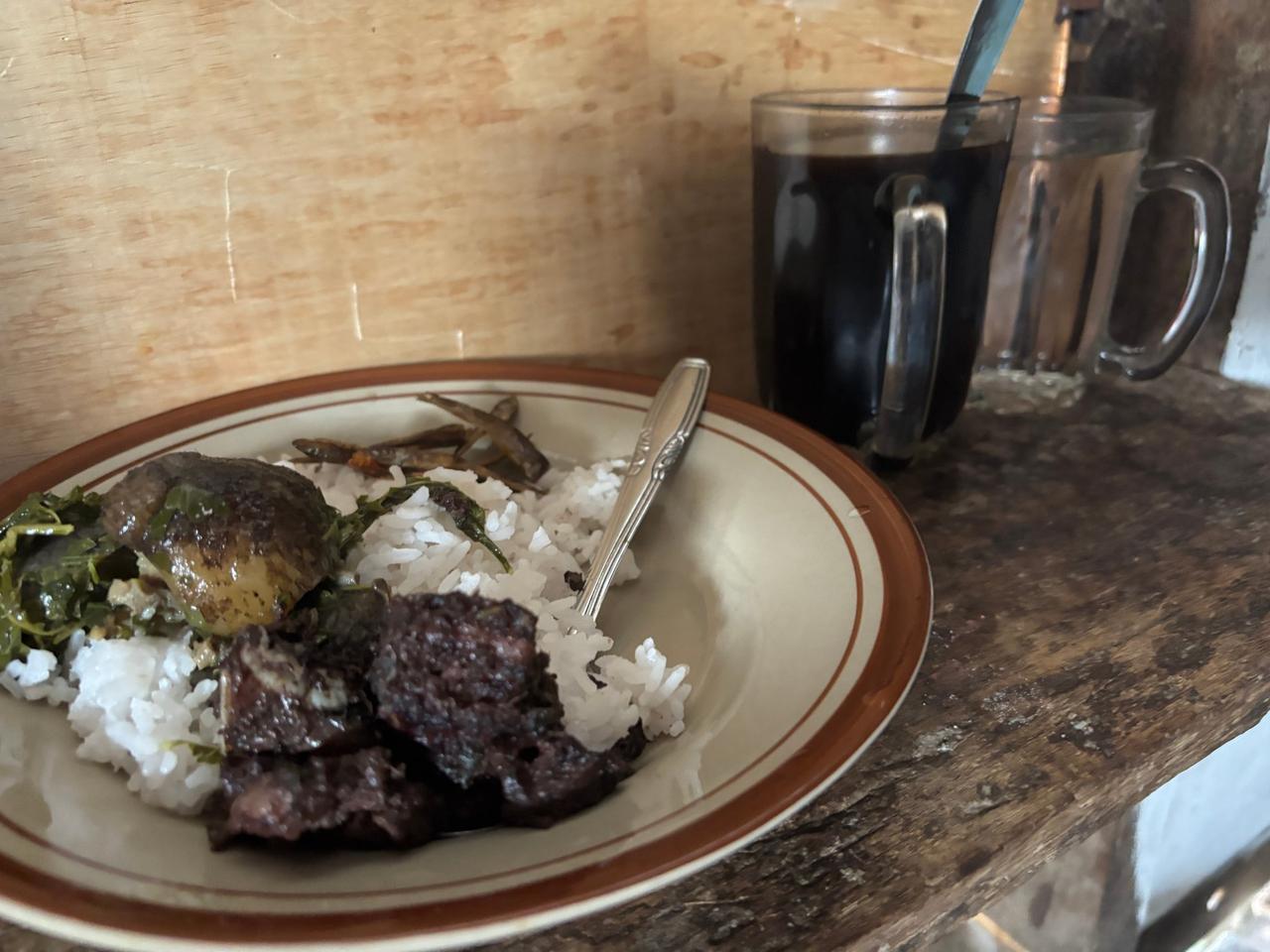 A plate of rice with pork, greens, and small fried fish beside a cup of black coffee