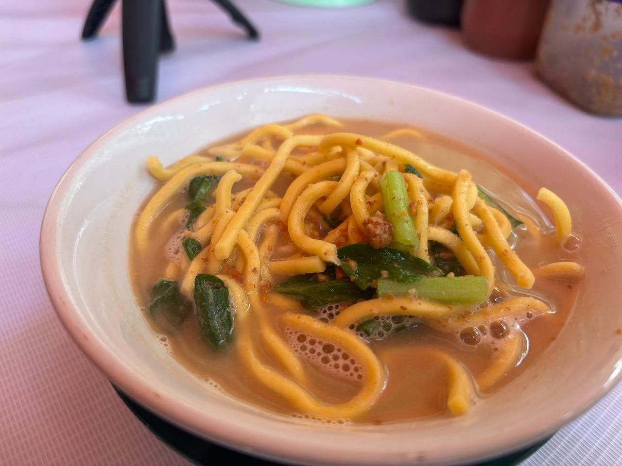 A bowl of noodle soup with greens and chili on a table