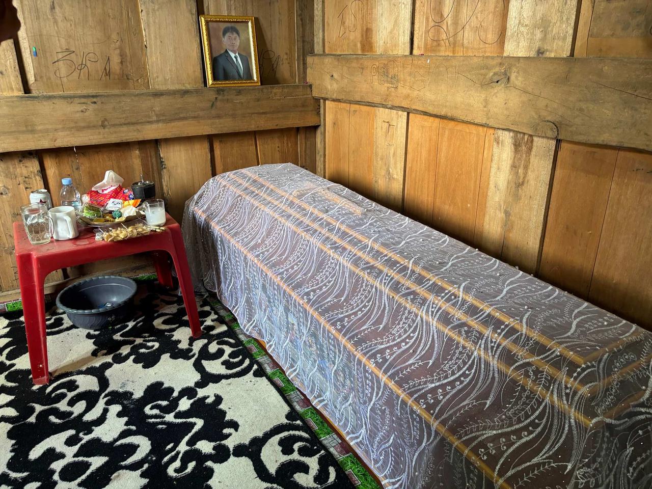 A sunlit wooden room with a shrouded coffin and a framed portrait of the deceased resting on a shelf