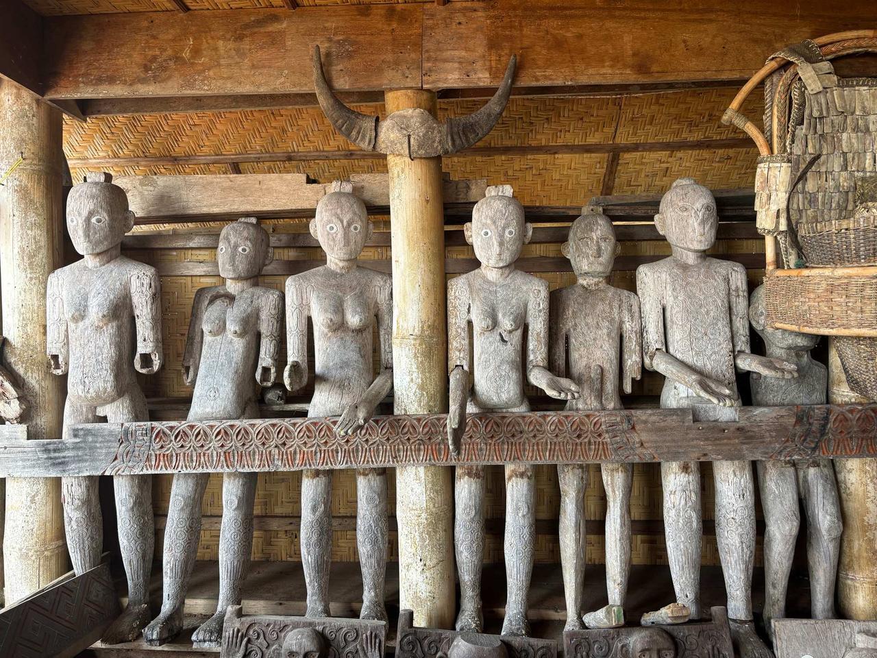 A row of carved wooden Tau Tau effigies standing beneath a buffalo-horn ornament inside a traditional Torajan structure