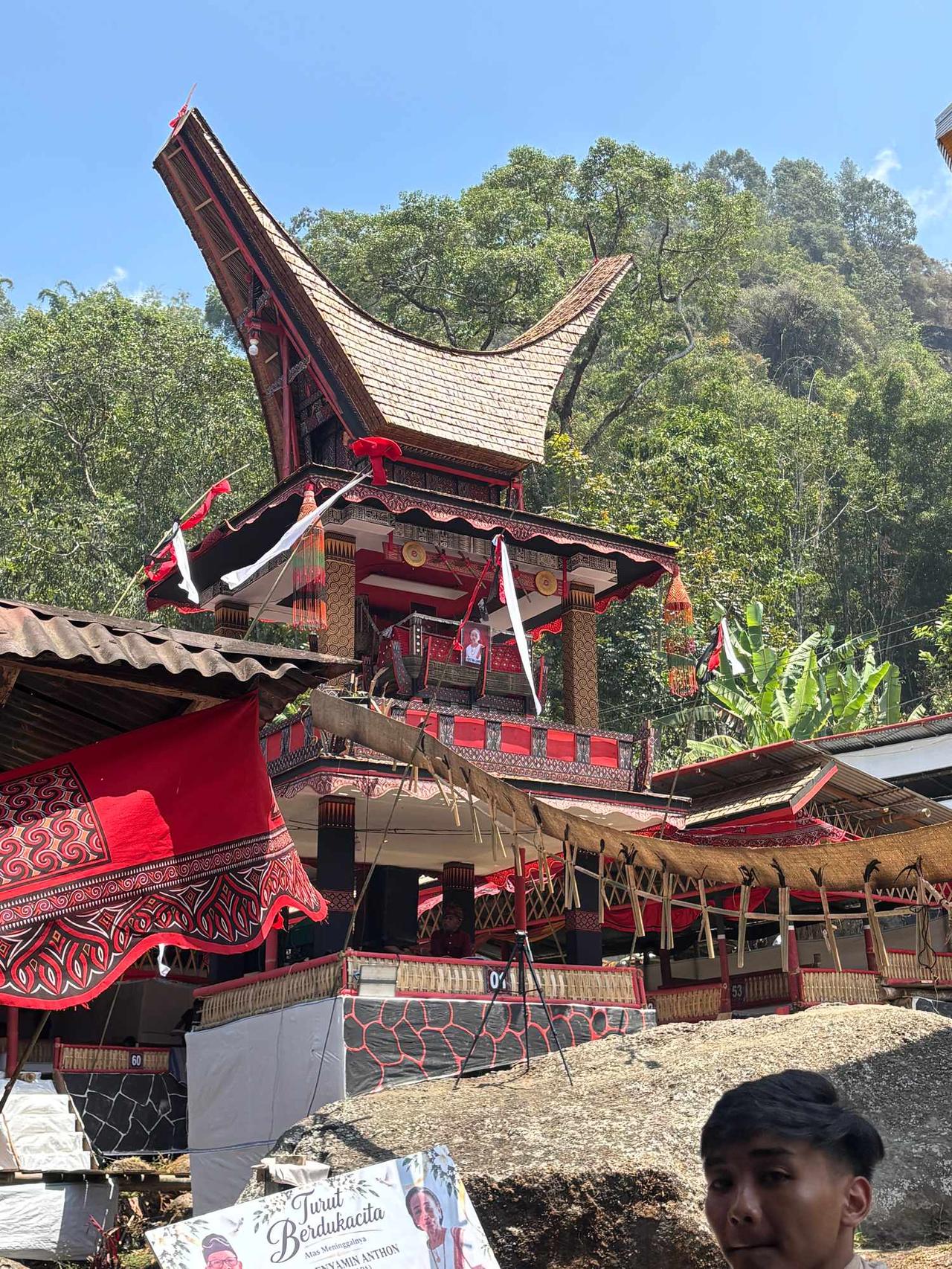 The towering red-and-black Lakian structure decorated with white banners and a portrait of the deceased, set against the forested hills