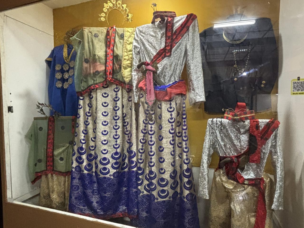 Busana istana kerajaan berwarna perak, biru, dan emas dipajang di balik kaca, dengan hiasan kepala terpasang di atasnya.