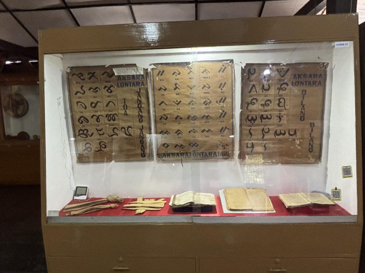 Bagan-bagan aksara Lontara kuno dan naskah tulisan tangan di atas kertas daun lontar, dipajang dalam lemari kaca.
